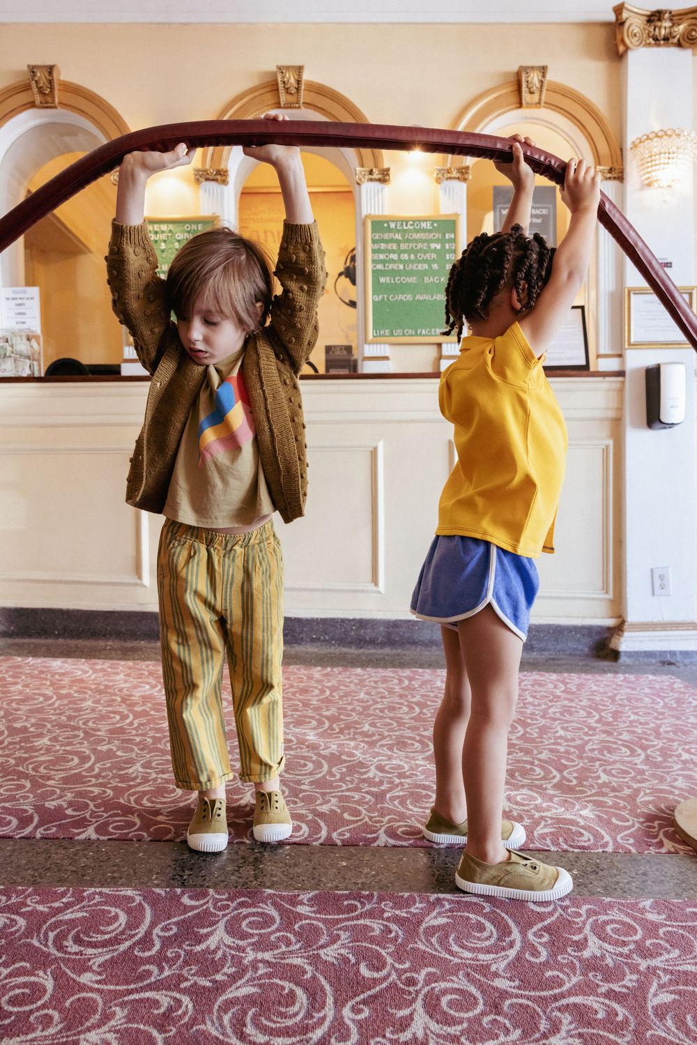 Two children playing on a rope barrier in an ornate movie theater lobby.