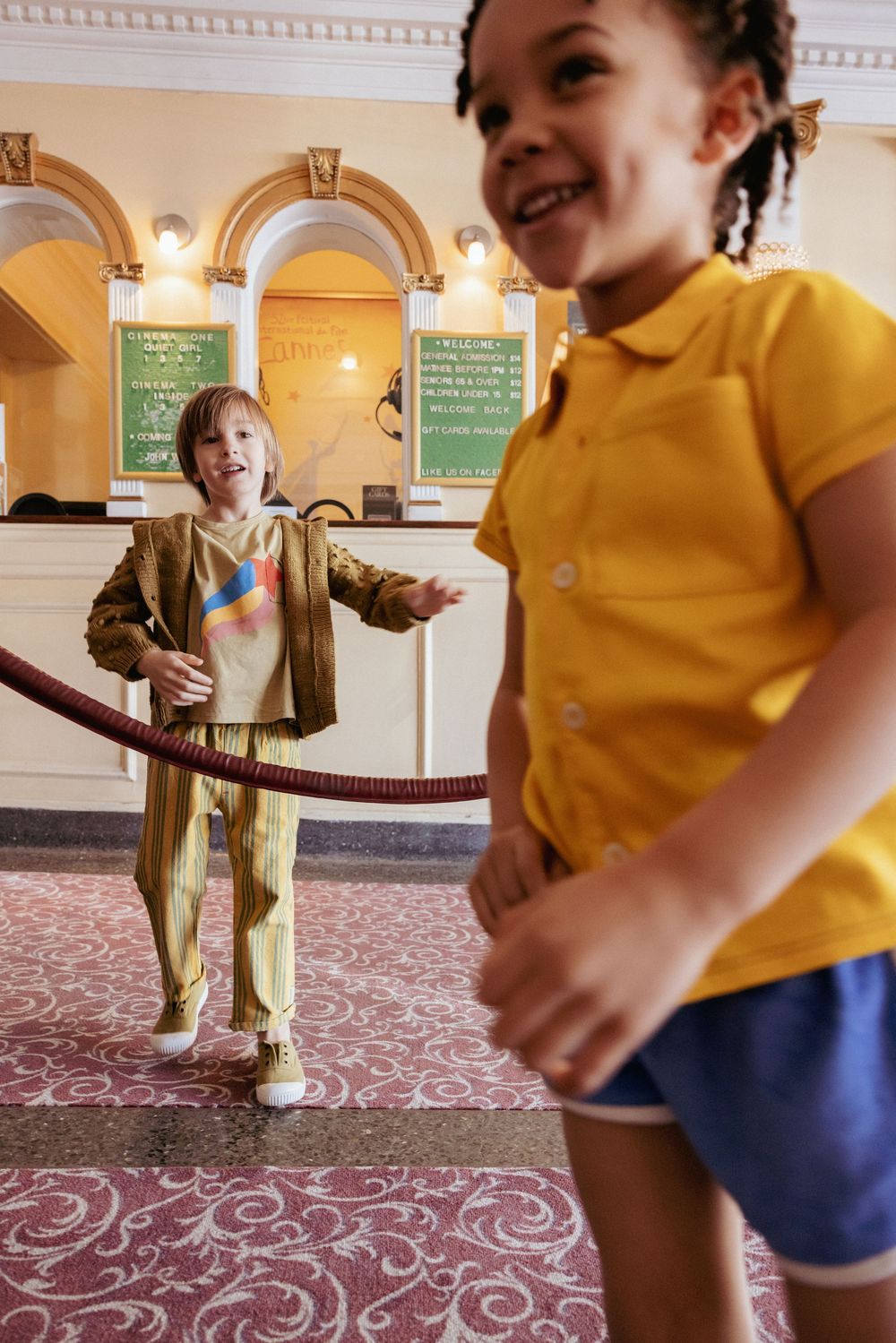 Two children laughing and playing with a velvet rope in a classic cinema lobby.