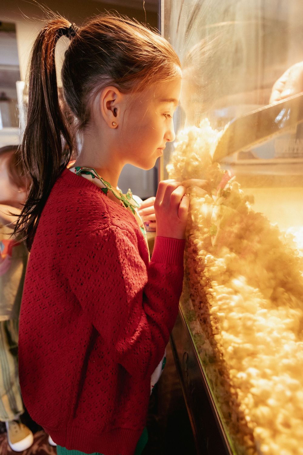 Young girl in red sweater gazes closely at illuminated popcorn display case at concession stand.