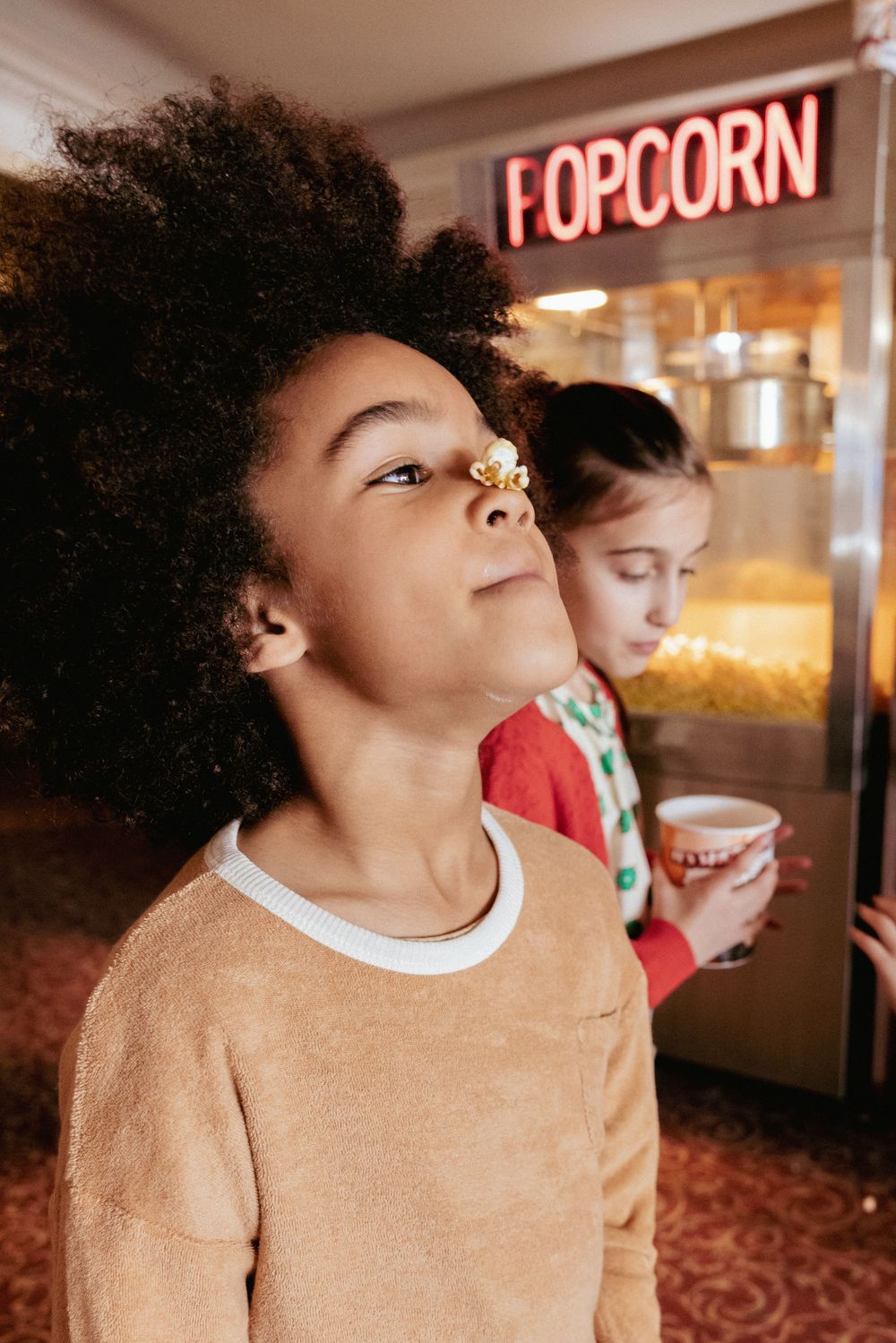 Young boy with popcorn on his nose standing near a popcorn machine at a cinema.