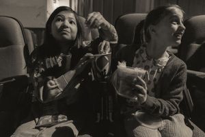 Black and white photo of two girls eating snacks together in movie theater seats.