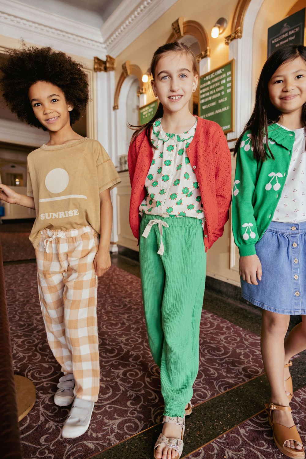 Three stylish girls smiling in a vintage movie theater lobby wearing colorful outfits.