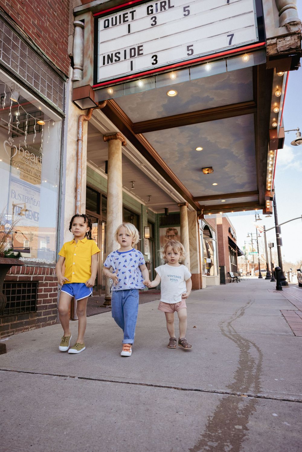 Three young children walking hand in hand outside a classic cinema theater marquee.