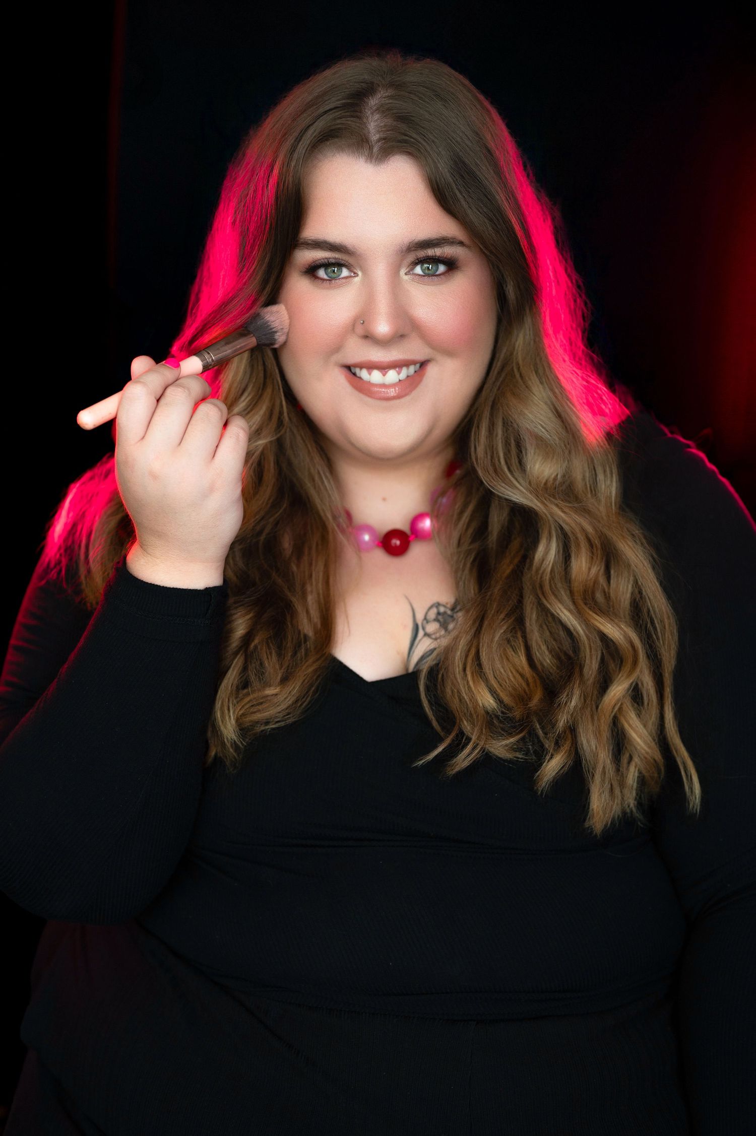 Woman in black top holding makeup brush, wearing pink necklace, with wavy hair and tattoo, posed against dark background.