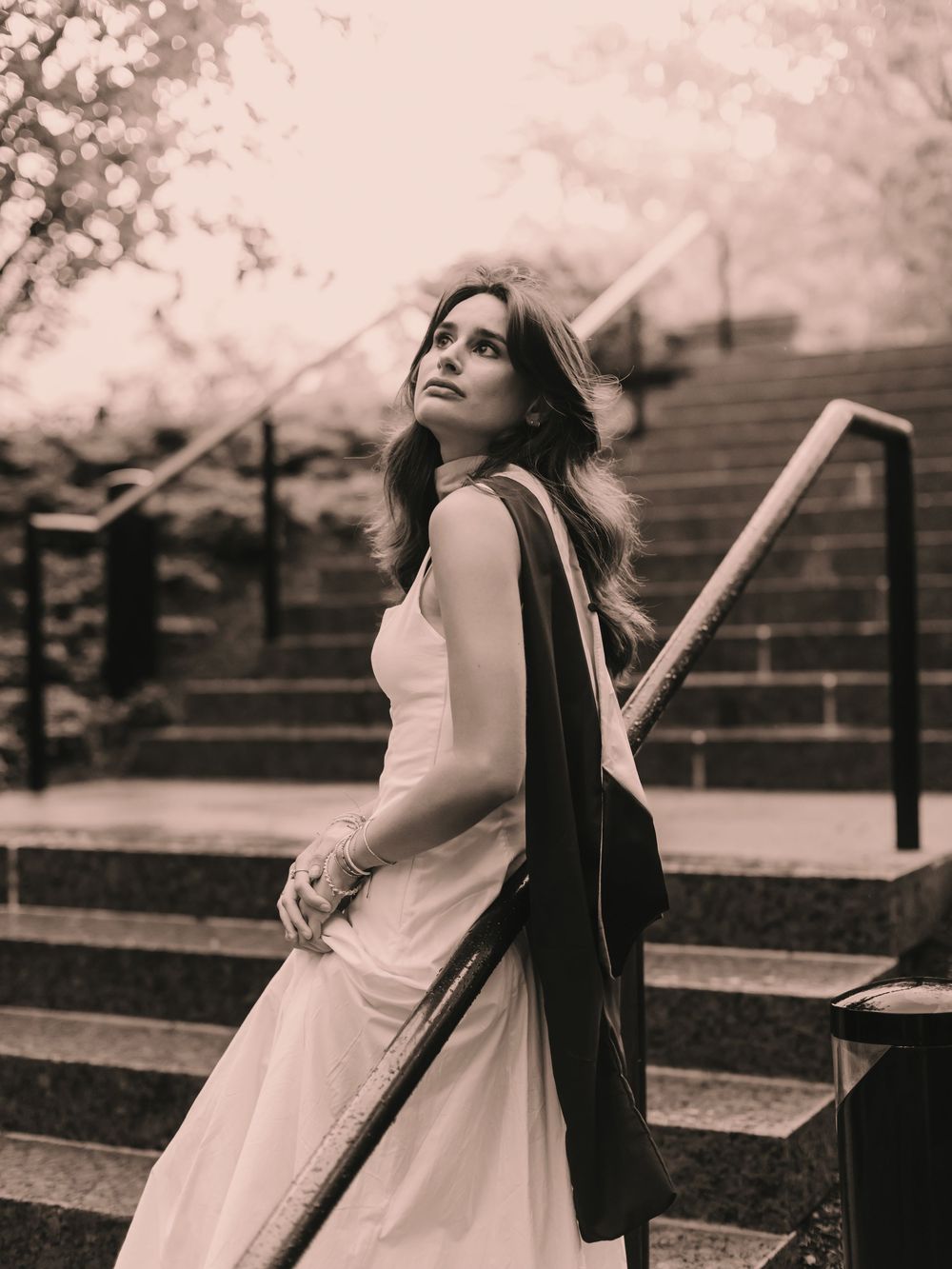 Candid photo of a Boston college graduate leaning against a railing on campus in a cinematic B&W setting wearing a graduation sash