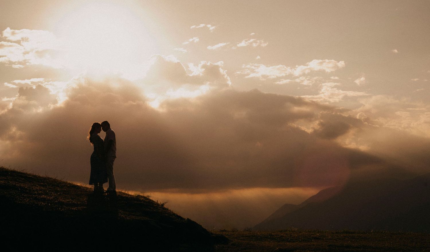 Silhouette of couple embracing on mountain hillside at golden sunset with dramatic clouds and rays of light.