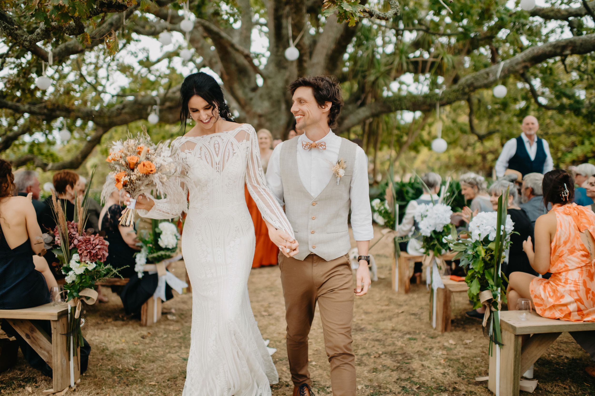 Bride and groom walk joyfully down outdoor wedding aisle under large oak tree, guests seated on wooden benches.