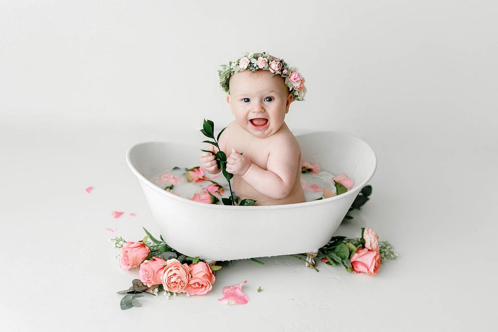 Smiling baby girl in floral crown sits in white bathtub surrounded by pink roses for a milk bath photo session.