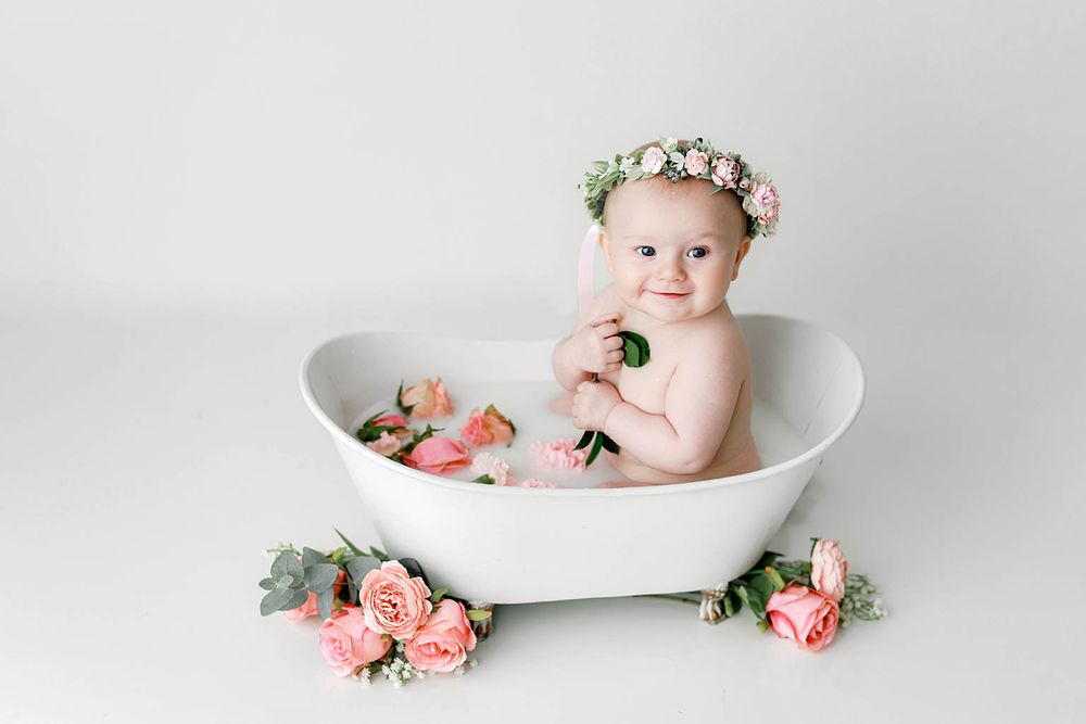 Baby girl with flower crown sits in white bathtub with pink roses during milestone photo session.