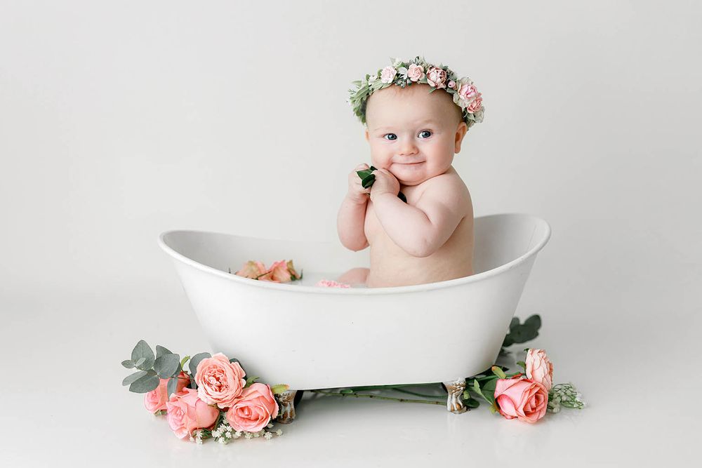 Adorable baby girl in floral crown sits in white clawfoot tub surrounded by pink roses.