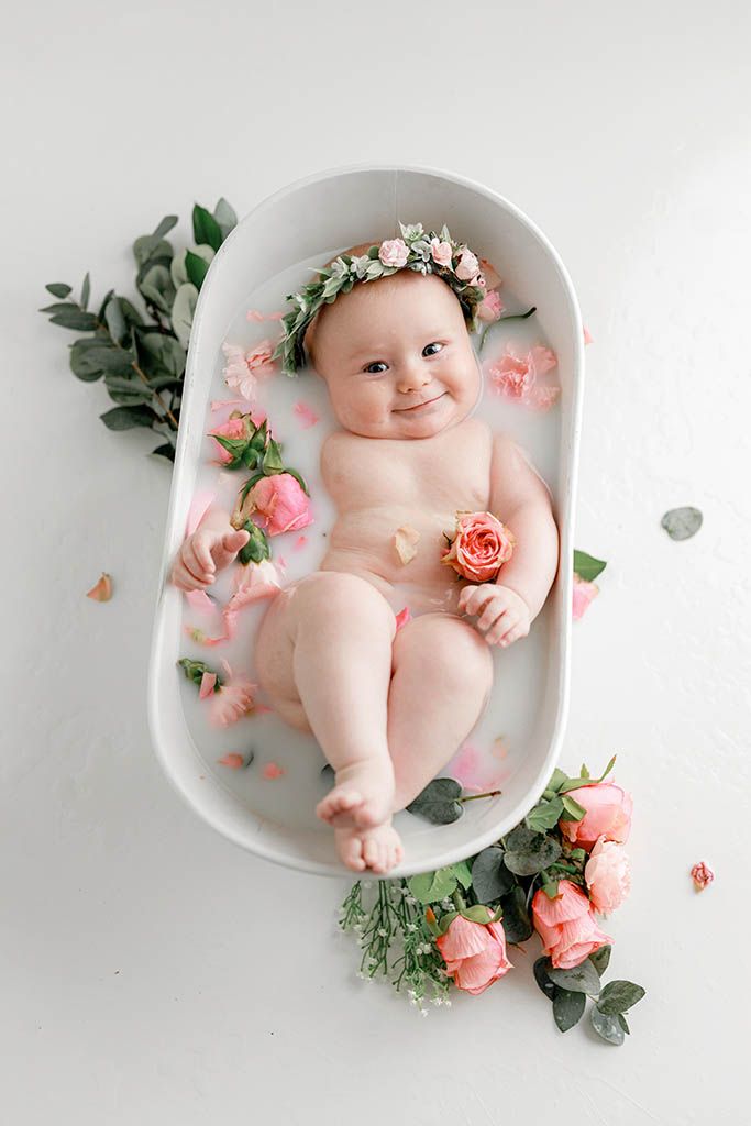 Adorable baby girl in floral crown smiling in white bathtub with pink roses and milk bath.