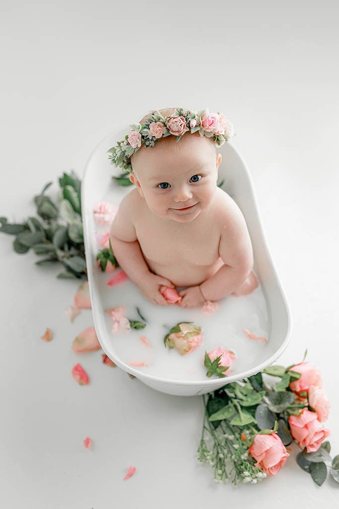 Baby girl wearing pink floral crown relaxes in white tub with milk bath and rose petals.