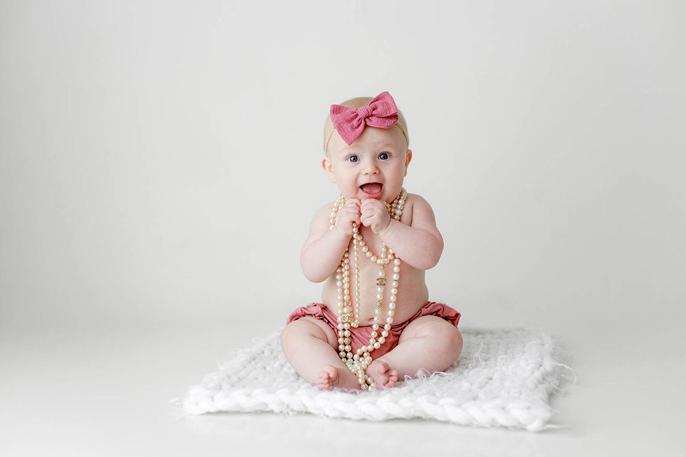 Cute baby girl sitting on white fur wearing pink bow headband and pearl necklace in studio portrait.