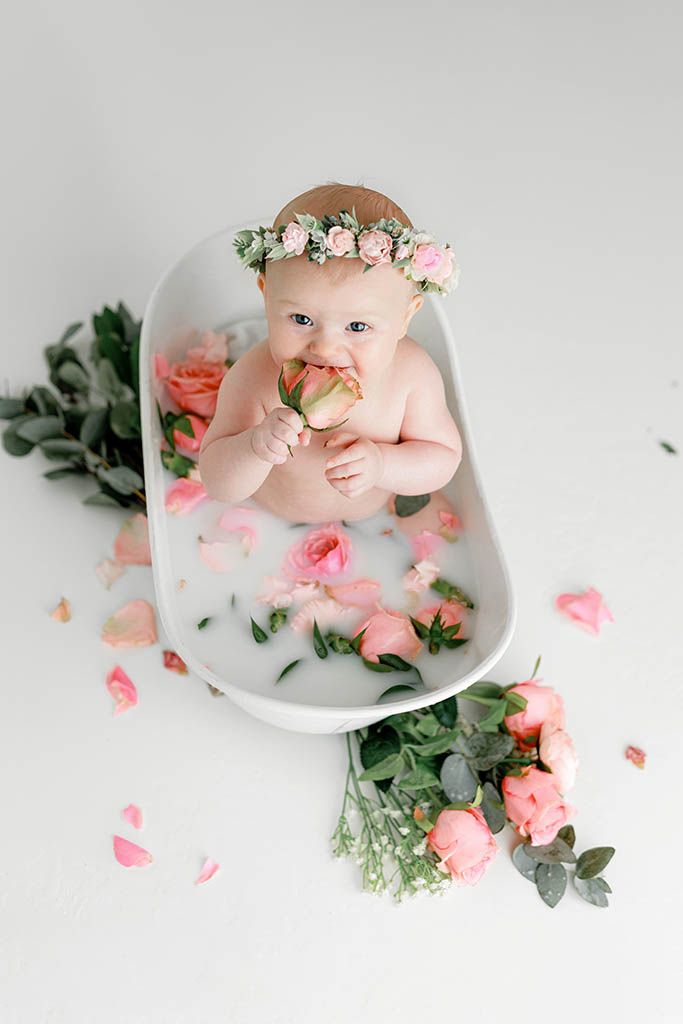 Baby girl wearing flower crown chewing on pink rose while sitting in milk bath with roses.