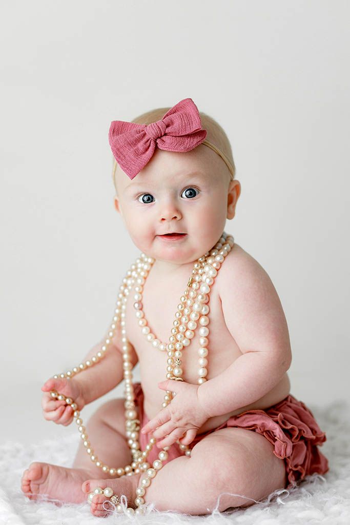 Adorable baby girl wearing pearl necklaces and a pink bow headband sitting on a white fluffy blanket.