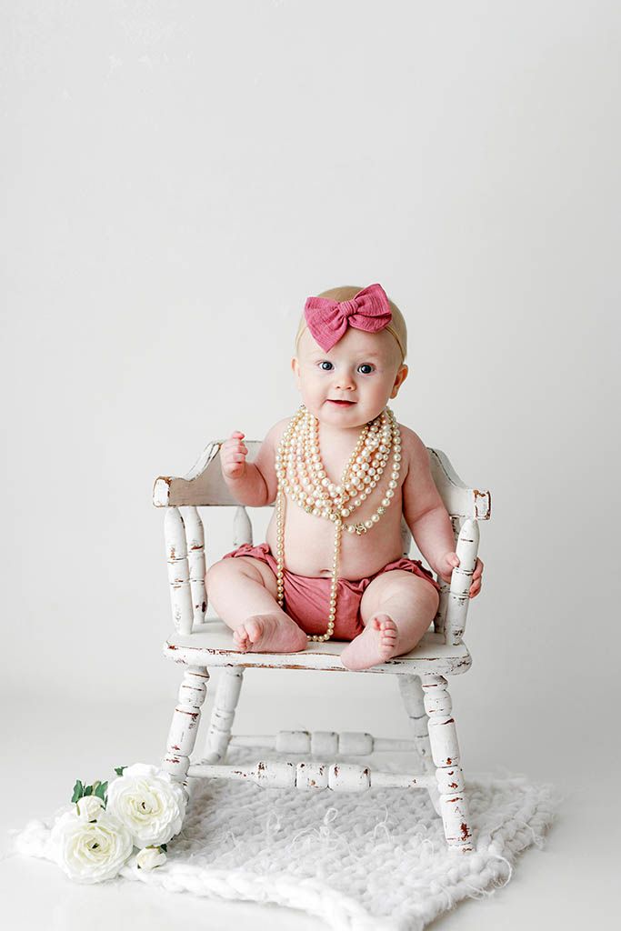 Baby girl in pink bloomers and pearl necklace sits on white rustic chair with bow headband.
