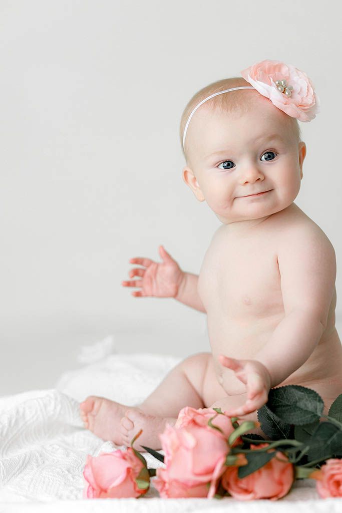 Adorable baby girl wearing a pink floral headband sitting with pink roses on a white background.