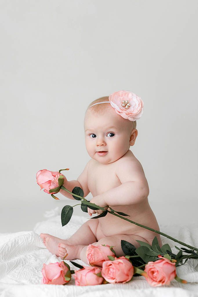 Adorable baby girl with pink flower headband sits holding pink roses on white backdrop in studio portrait.