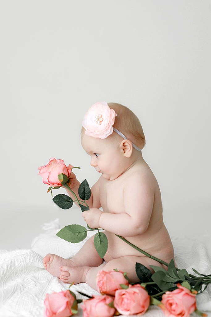 A baby girl wearing a floral headband sits on white fabric, holding a pink rose surrounded by roses.