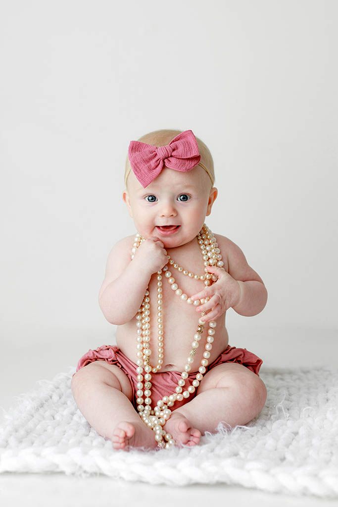 Adorable baby girl in pink bloomers and bow headband sitting on white rug playing with pearl necklaces.