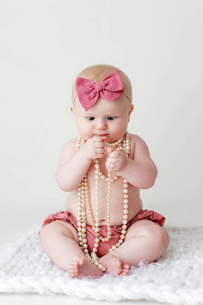 Baby girl in pink bloomers and bow headband playing with pearl necklaces, sitting on white fluffy blanket.