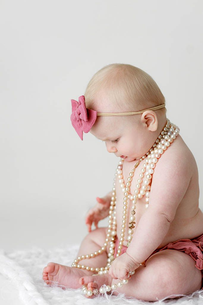 Adorable baby girl wearing pearl necklaces and pink bow headband sitting on white fur.