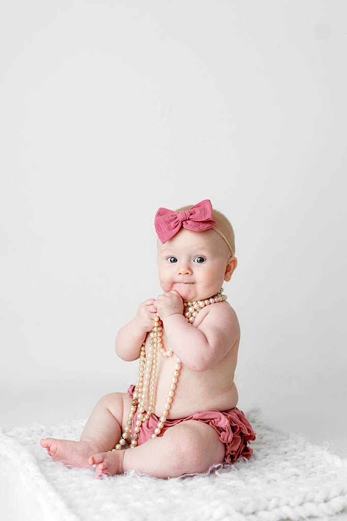 Cute baby girl in pink bow headband chews pearl necklace while sitting on white fluffy rug.