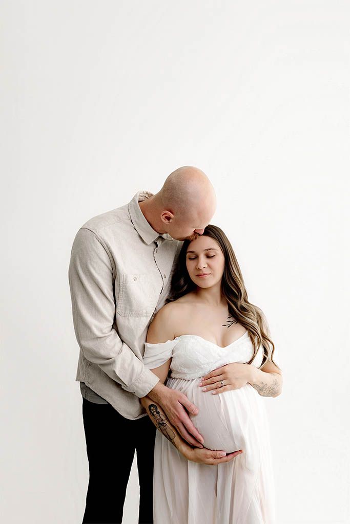 Couple sharing a tender maternity moment, man kissing woman's head while both cradle her pregnant belly in white studio.