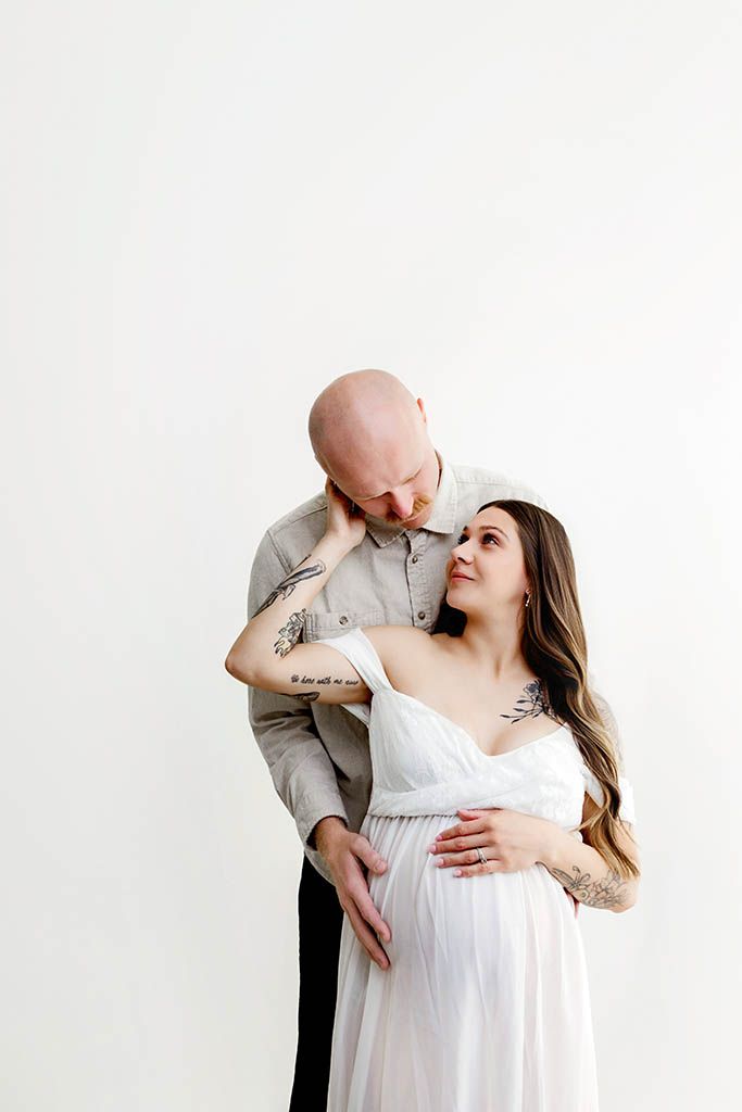 Couple sharing a tender maternity moment, tattooed man embracing pregnant woman in white dress against white background.