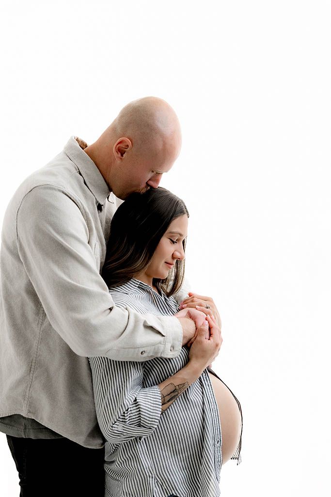 A bald man tenderly embraces a pregnant woman from behind, both looking down at her baby bump in a white studio.