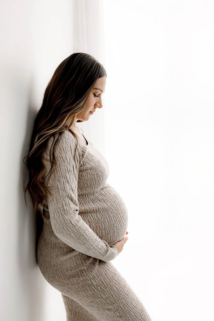 Pregnant woman in gray knit dress cradling baby bump against white wall, smiling downward in a serene maternity photo.