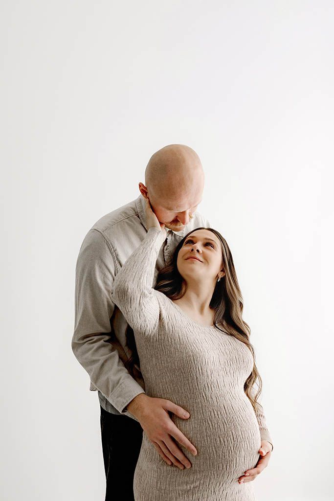 A bald man tenderly kisses a pregnant woman's forehead as she gazes up at him, both wearing neutral tones.