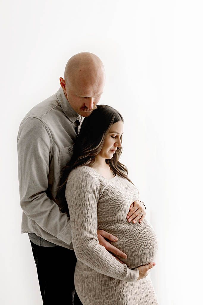 A couple tenderly cradles a pregnant belly during a maternity photo session against a white background.