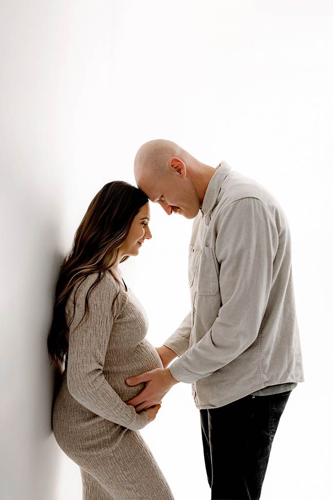 Couple sharing a tender maternity moment, hands on pregnant belly, leaning foreheads together against a white background.