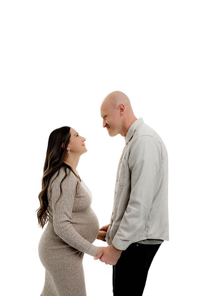 Pregnant couple holding hands and smiling at each other in a maternity photo shoot against a white background.