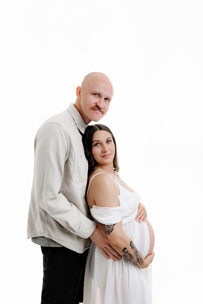 A bald man with a mustache embraces a pregnant woman in a white dress against a white background.