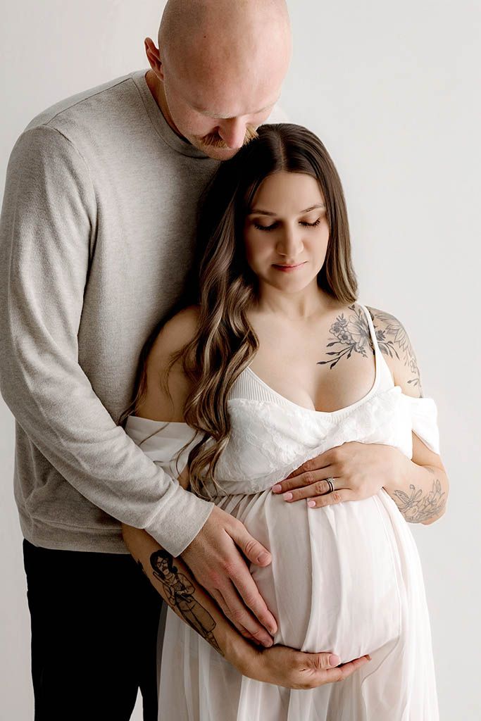 A tattooed couple embraces while cradling a pregnant belly in a soft white maternity portrait.