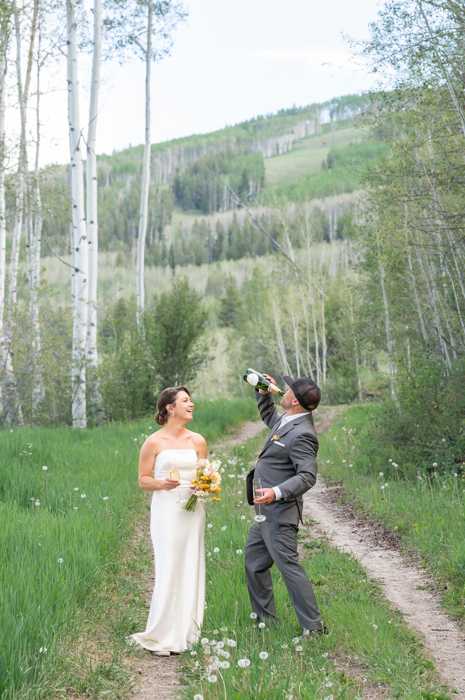 Bride and groom celebrate outdoors among aspen trees, groom drinking from champagne bottle on a scenic mountain path in Beaver Creek