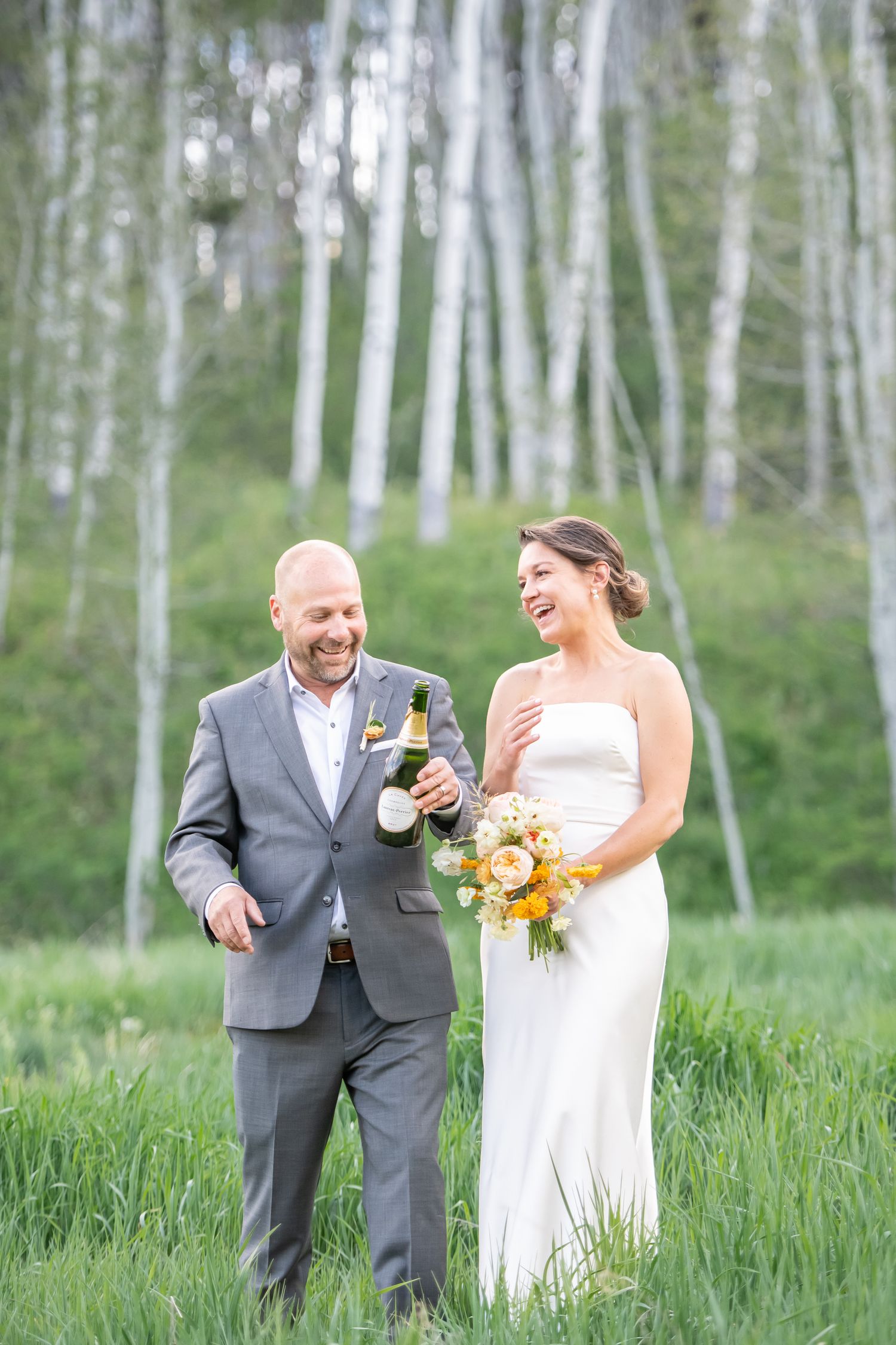 Laughing bride and groom celebrate with champagne in a lush green meadow surrounded by aspen trees in Beaver Creek Colorado