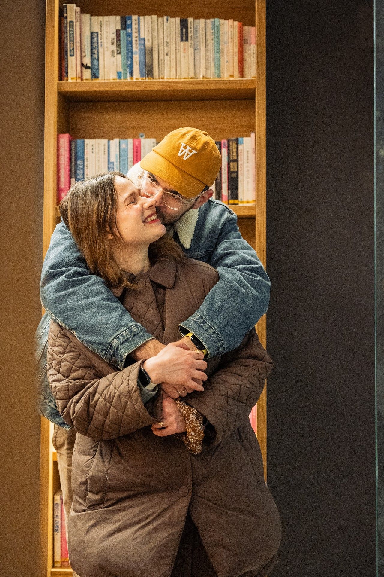 A couple embracing and smiling in front of a wooden bookshelf, woman in brown quilted coat, man in orange cap.