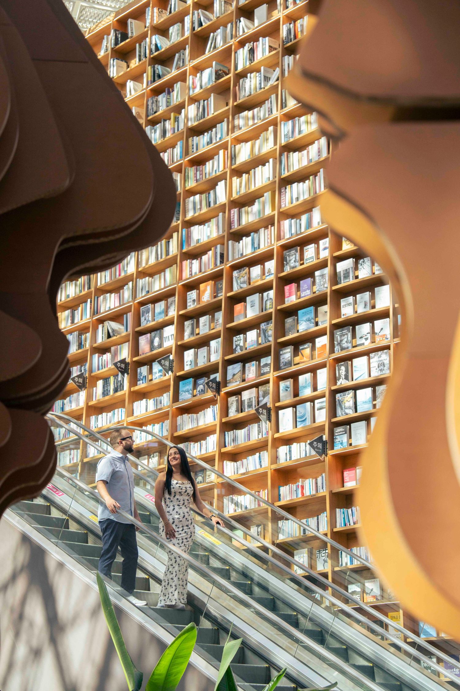 Couple descending escalator in stunning library with floor-to-ceiling bookshelves and artistic sculptural frames in foreground.
