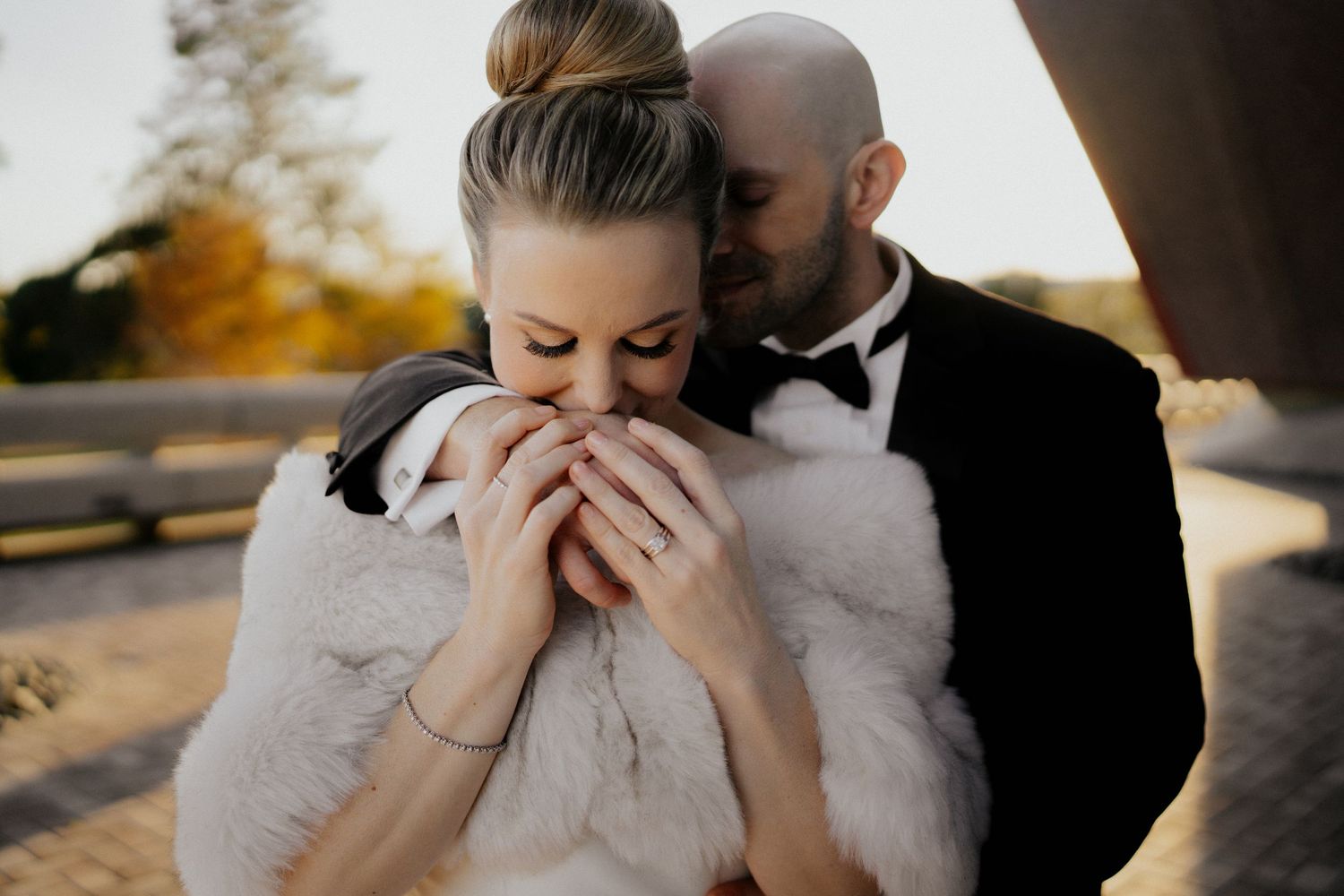 Bride in white fur wrap embraced by groom in black tuxedo during golden hour outdoor wedding portrait.