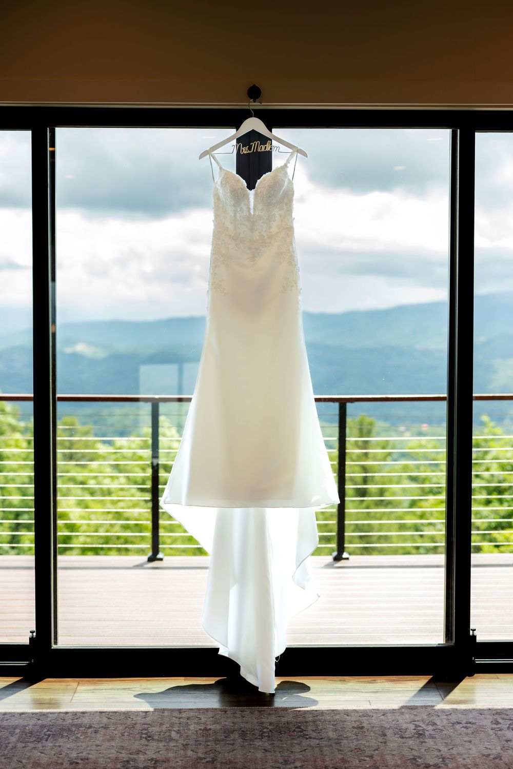 Elegant white lace wedding dress hanging on a hanger in front of large windows with mountain views.