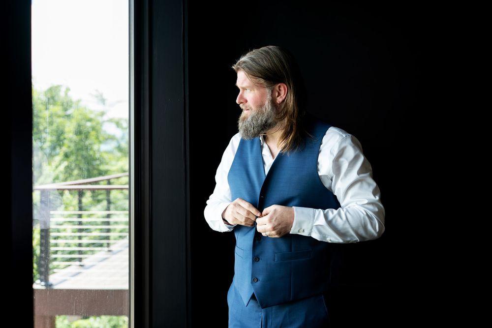 Bearded groom in blue vest and white shirt buttoning his jacket by a window on his wedding day.