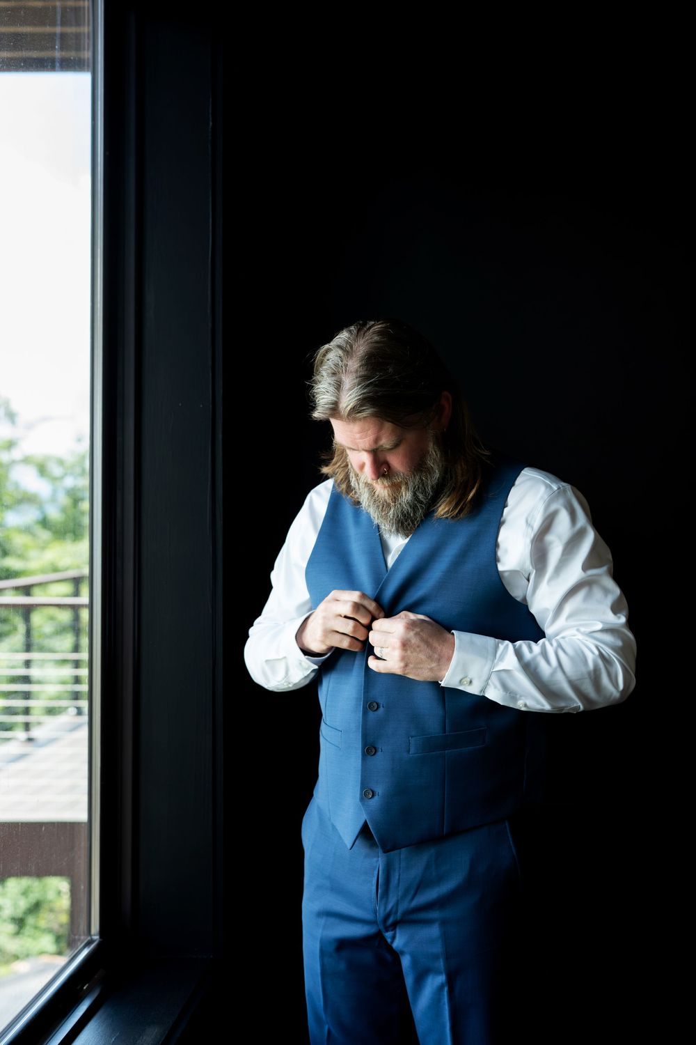 Bearded man in blue vest and white shirt buttoning up near a dark wall with natural window light.