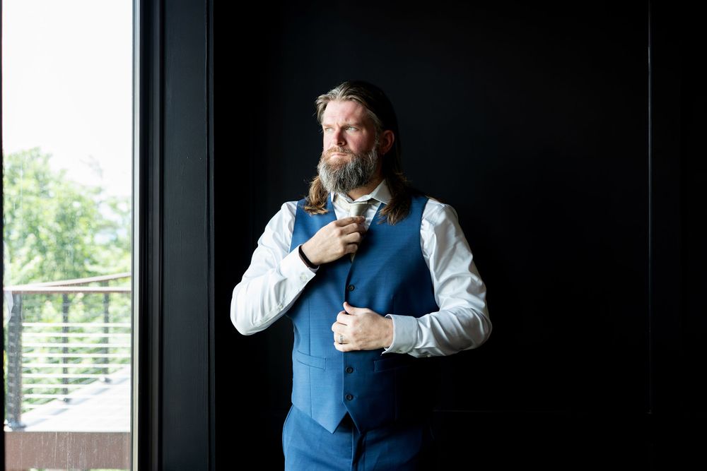 Bearded groom in blue suit vest adjusting his tie near a dark wall with natural light behind him.
