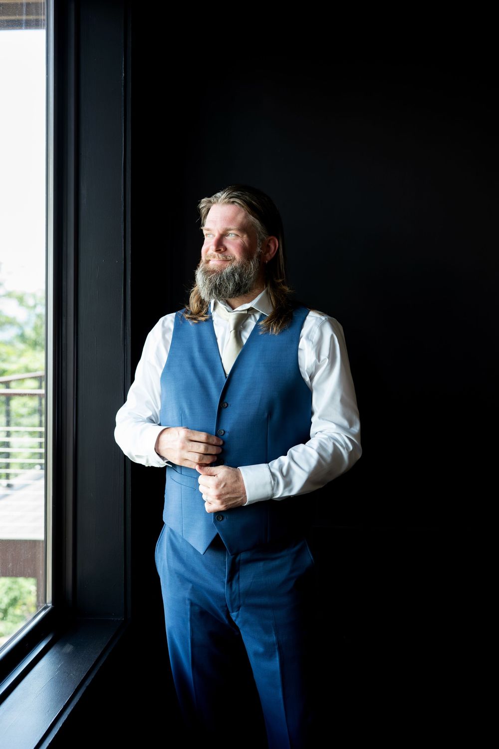 Bearded groom in blue vest and white shirt stands by window adjusting suit buttons before wedding ceremony.
