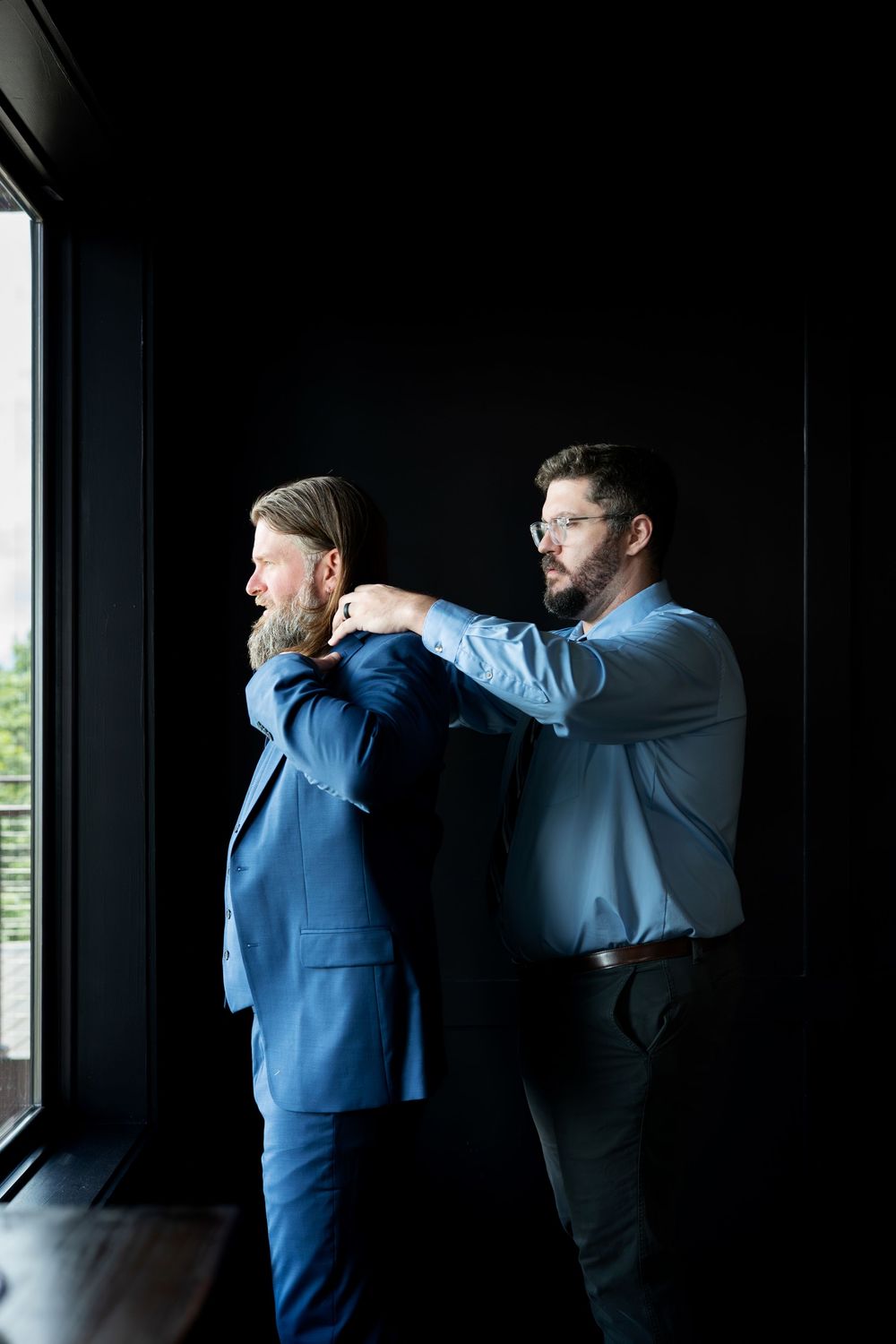Man in glasses helping another man in a blue suit jacket near a window in a dark room.