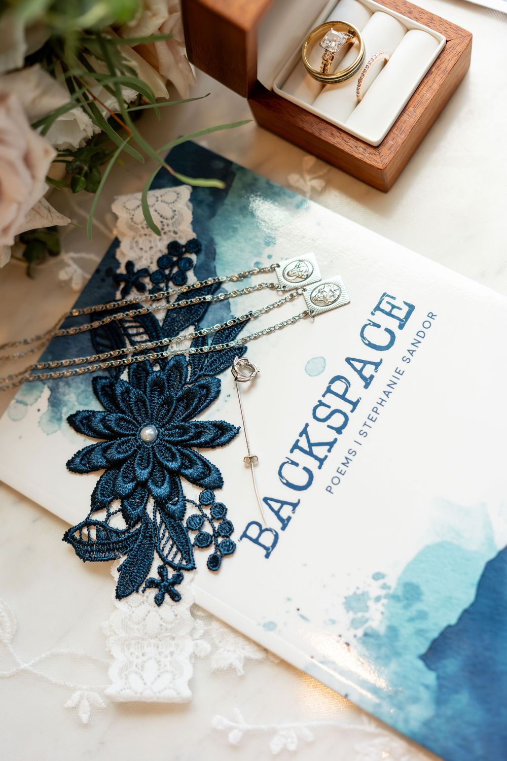 Blue floral lace wedding garter on Backspace poetry book with gold ring in box and delicate chain necklaces.