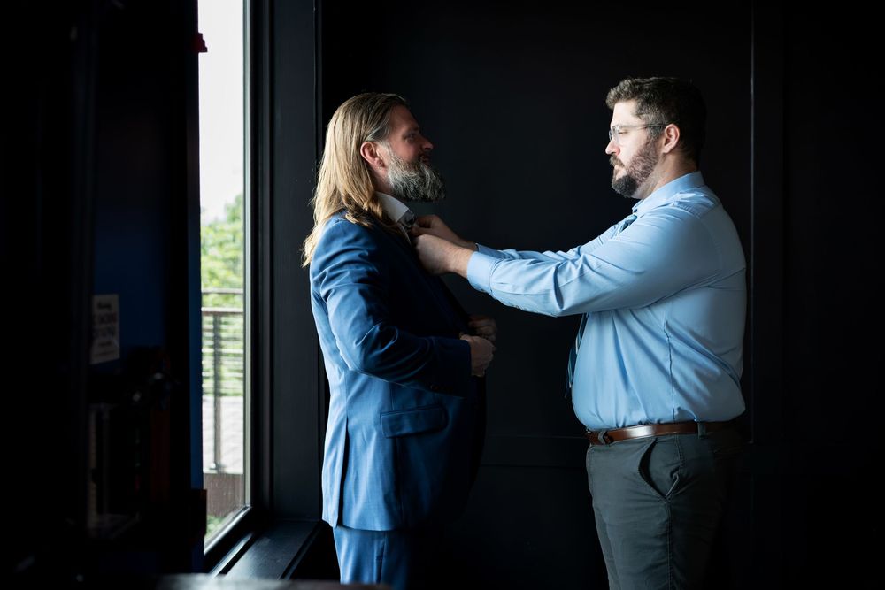 Two bearded men in formal attire adjust each other's suits indoors near a window in soft natural light.